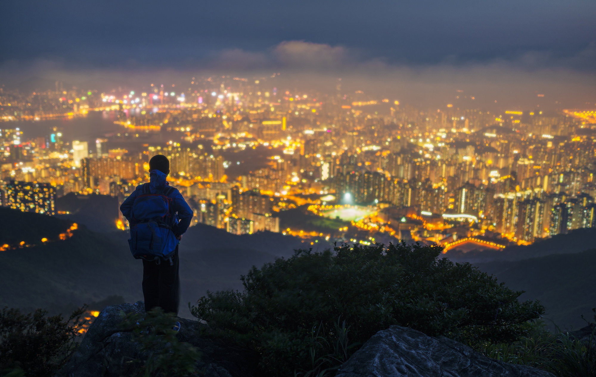 Hiker standing on a mountain peak overlooking Hong Kong skyline at night